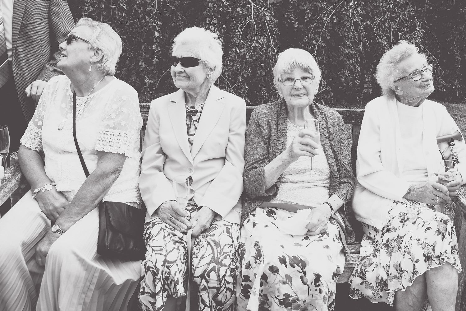 Four elderly women in floral skirts sitting on a bench laughing and enjoying themselves.