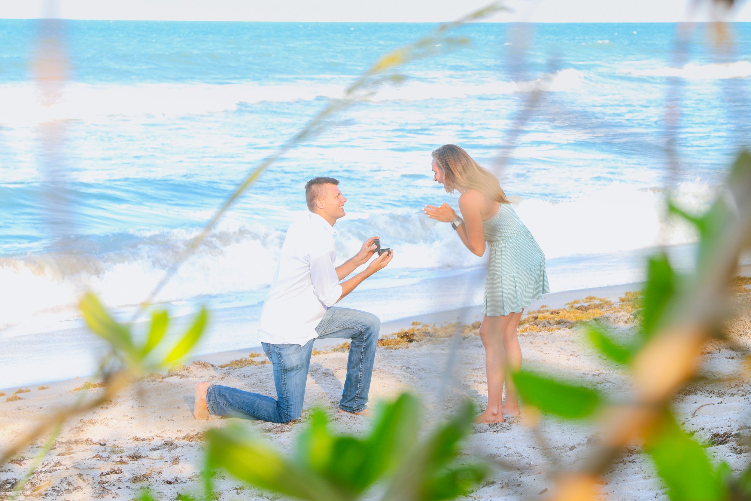 Man proposes to woman on a tropical beach, kneeling with a ring box as waves crash in the background.