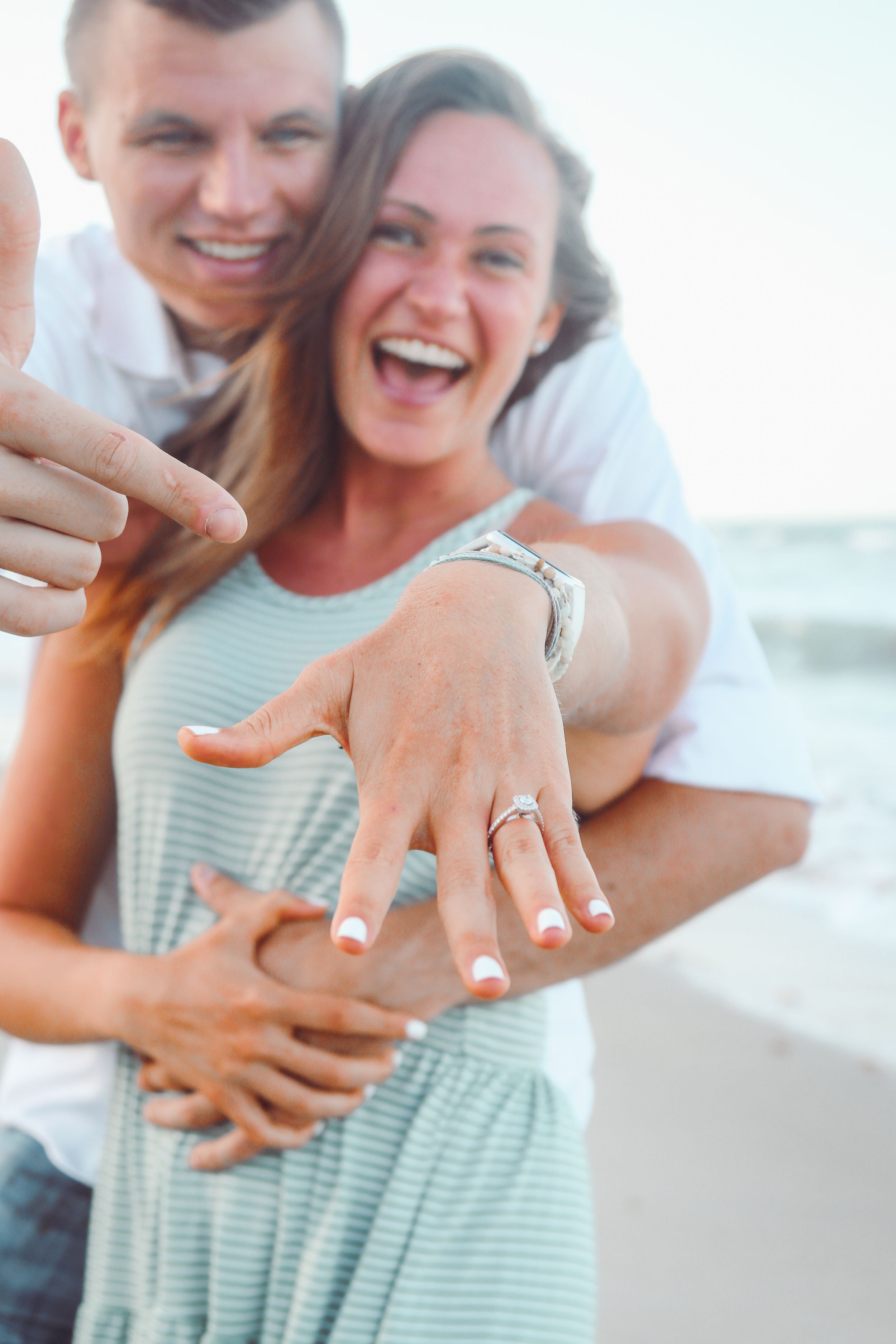Happy couple on beach showing off engagement ring, woman laughing with diamond ring on finger, man pointing at it.