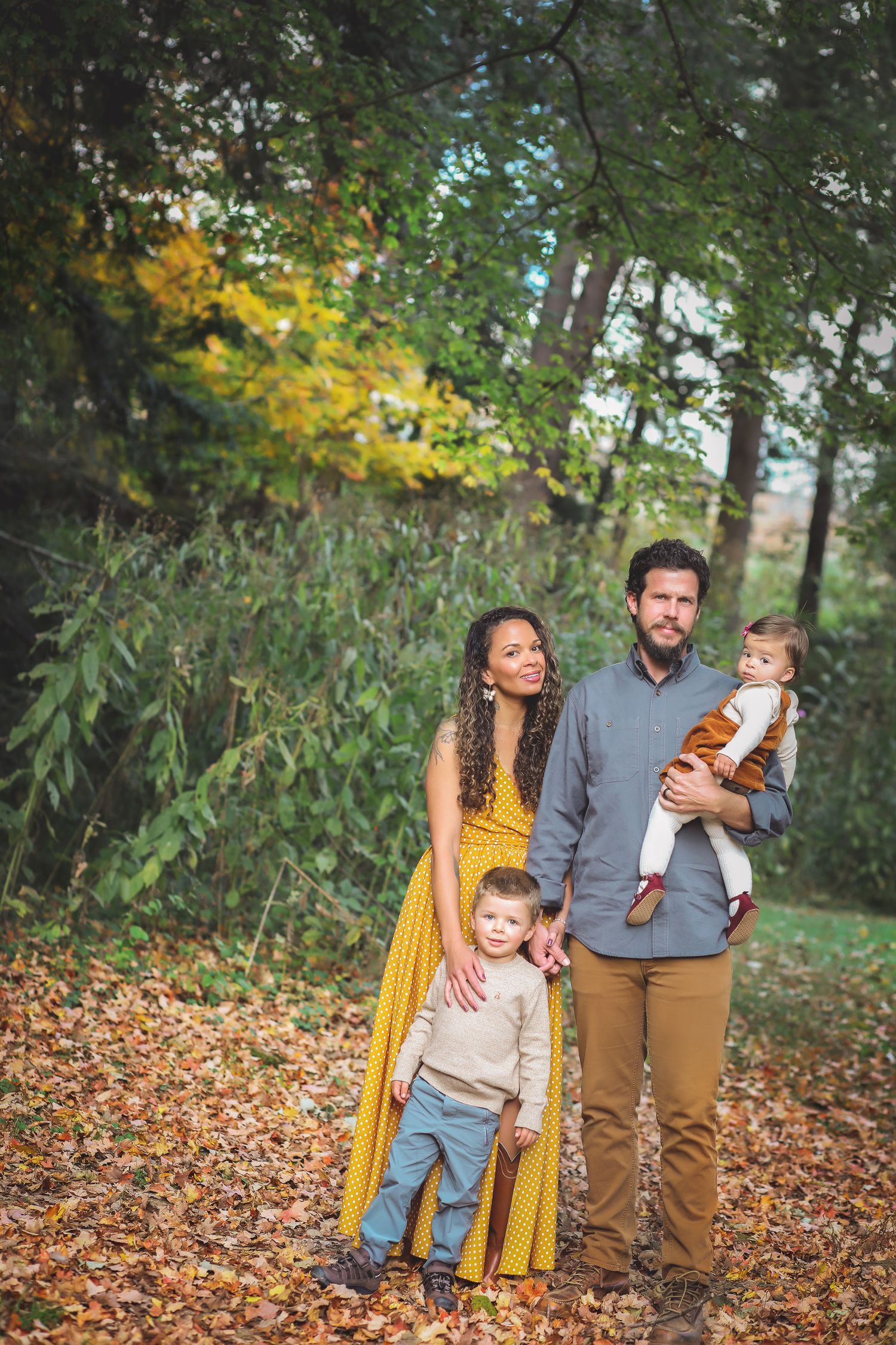 A fall family portrait in a wooded setting with autumn leaves and yellow dress details.
