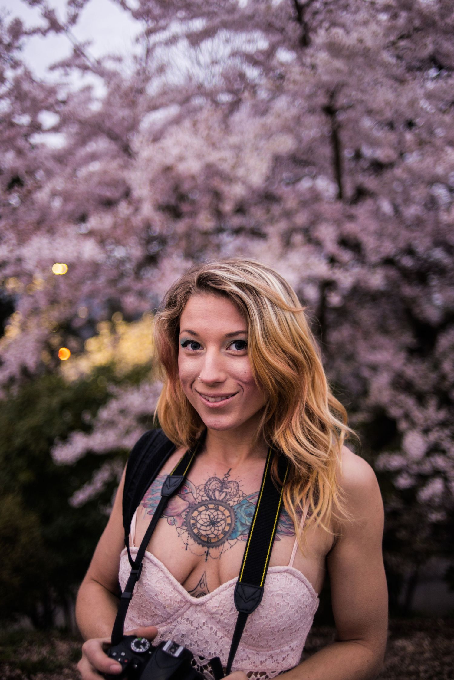 A photographer with a chest tattoo poses with camera gear against blooming cherry blossoms.