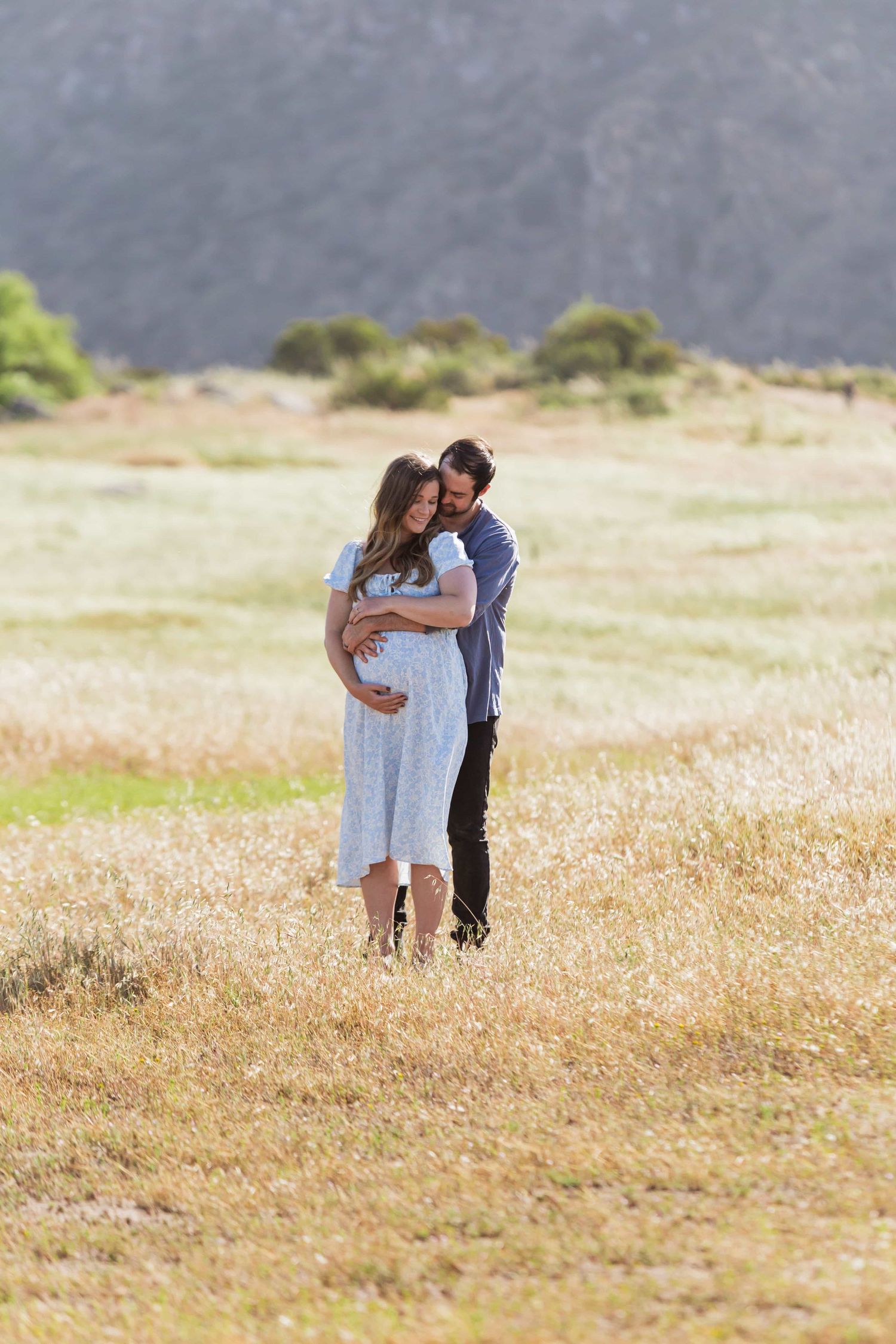 Couple embracing in a golden field, woman in white dress with visible baby bump, mountains behind.