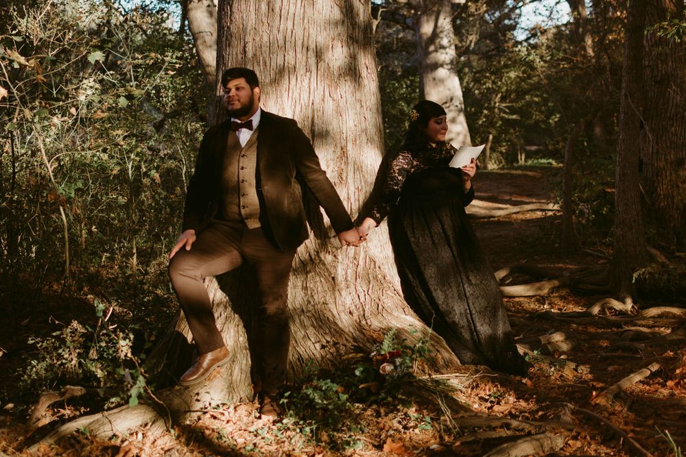 Two people pose playfully against a massive tree trunk in an autumn forest setting.