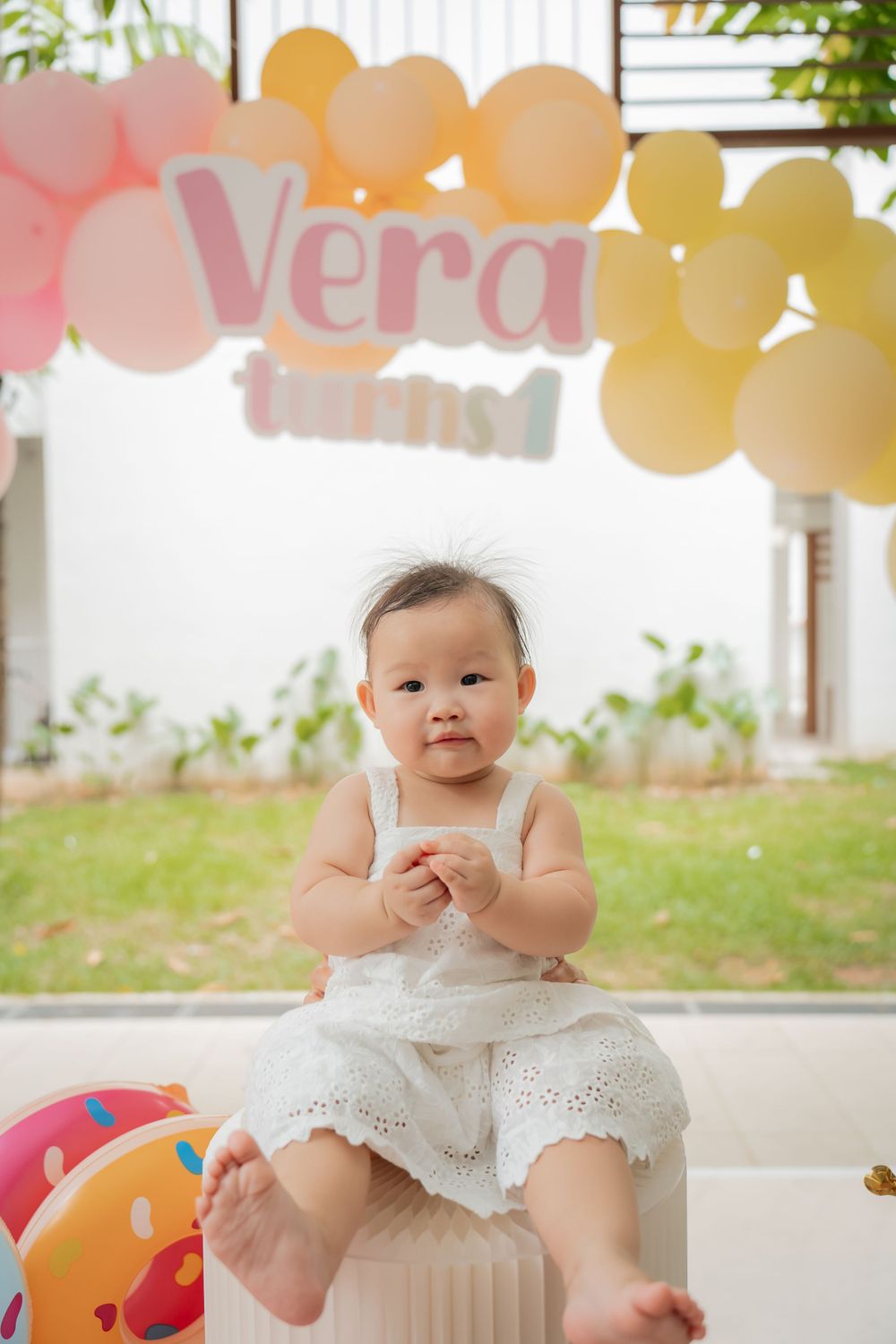 A baby in white attire sits under balloon decorations spelling out Vera.