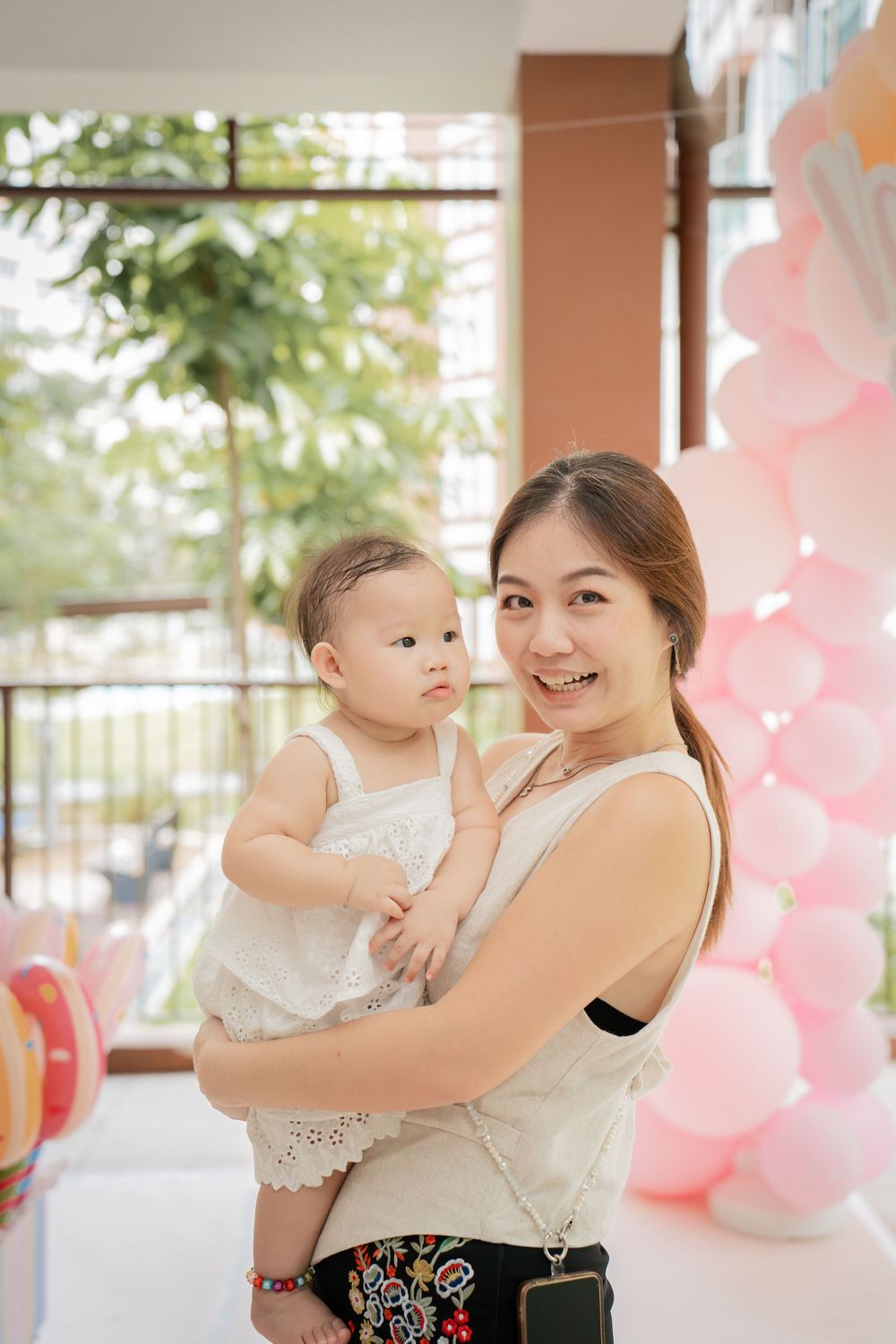 A baby in white dress at a celebration with pink balloon decorations in the background.