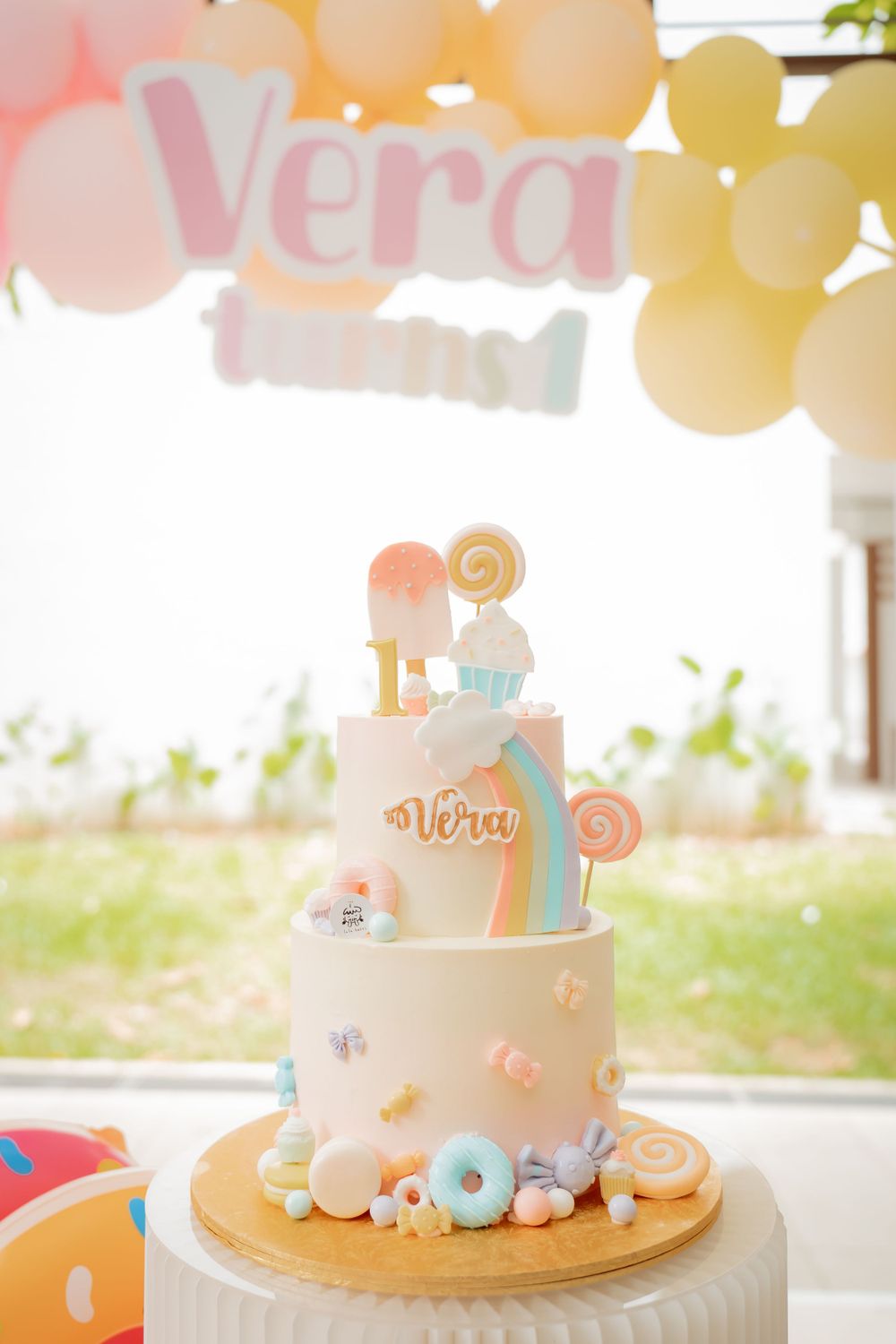 A pastel-colored celebration cake with lollipop decorations and the name Vera on display.