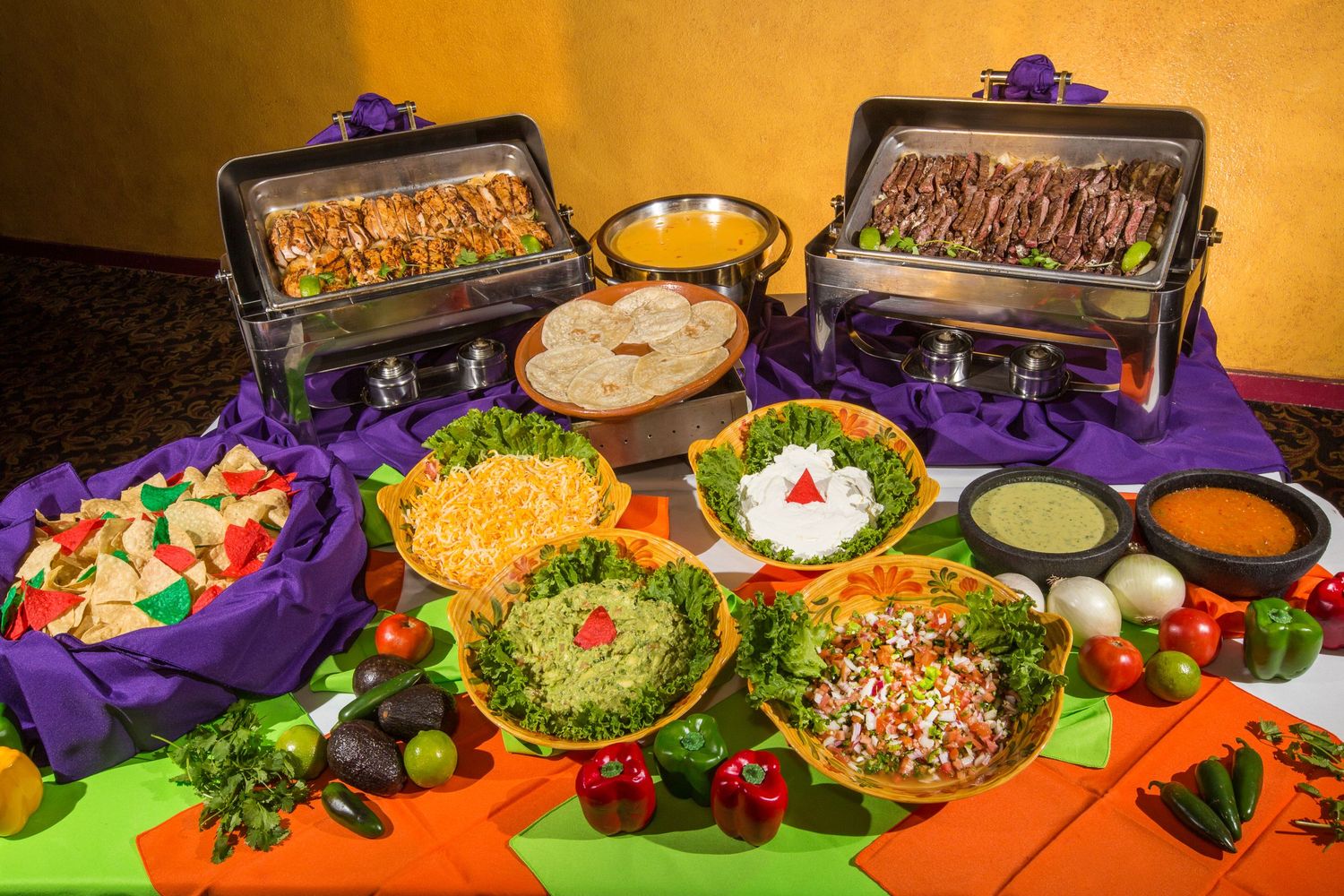 Mexican buffet spread with chafing dishes, colorful salads, and various dips on decorated table.