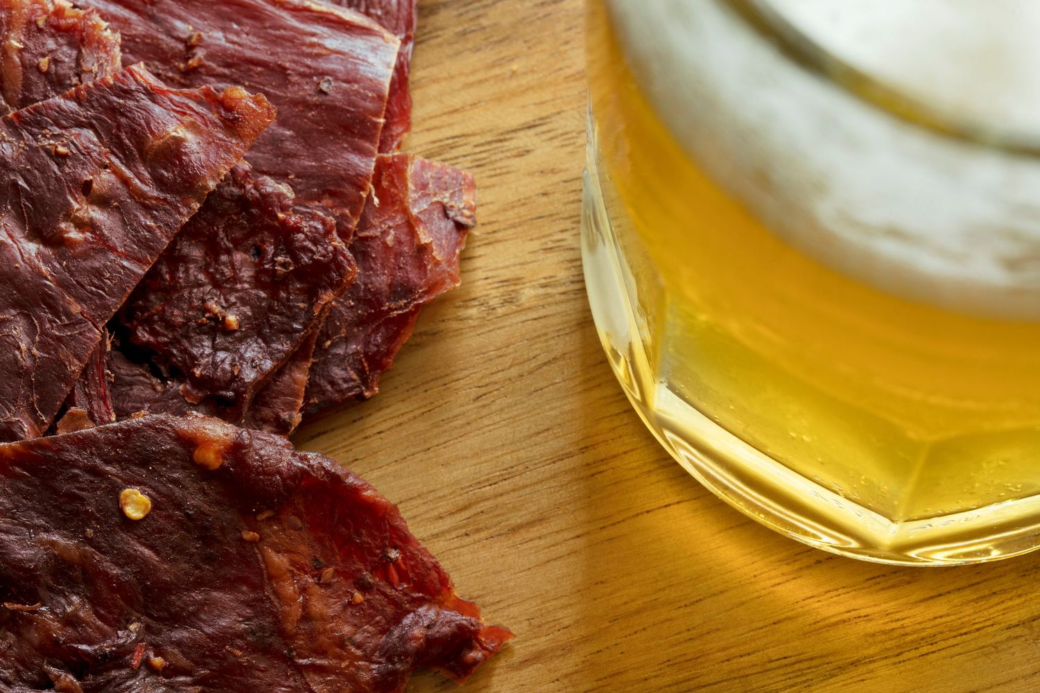 Pieces of dark beef jerky next to a glass of light beer on a wooden cutting board.