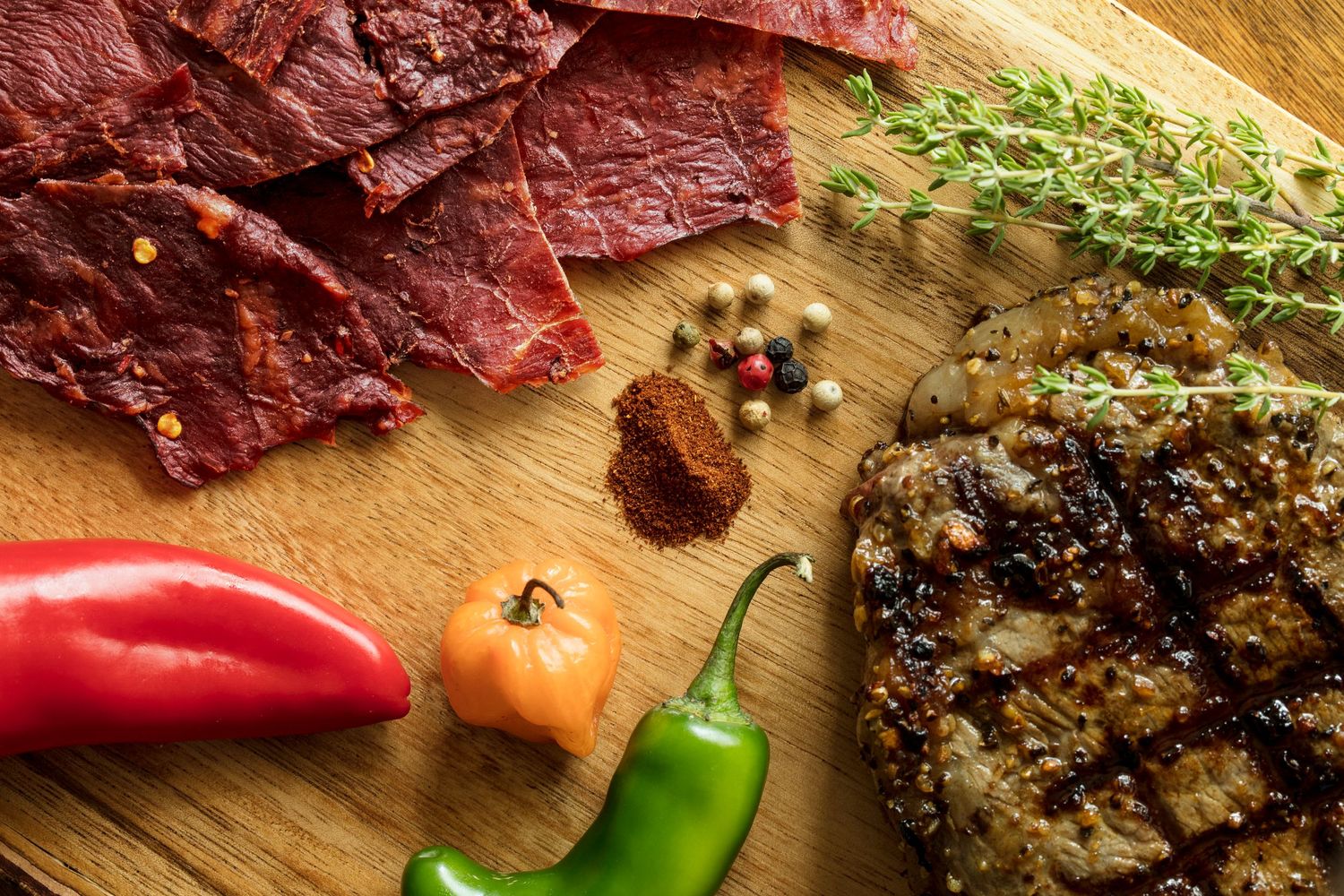 Assorted beef jerky with fresh peppers and spices on a wooden cutting board.