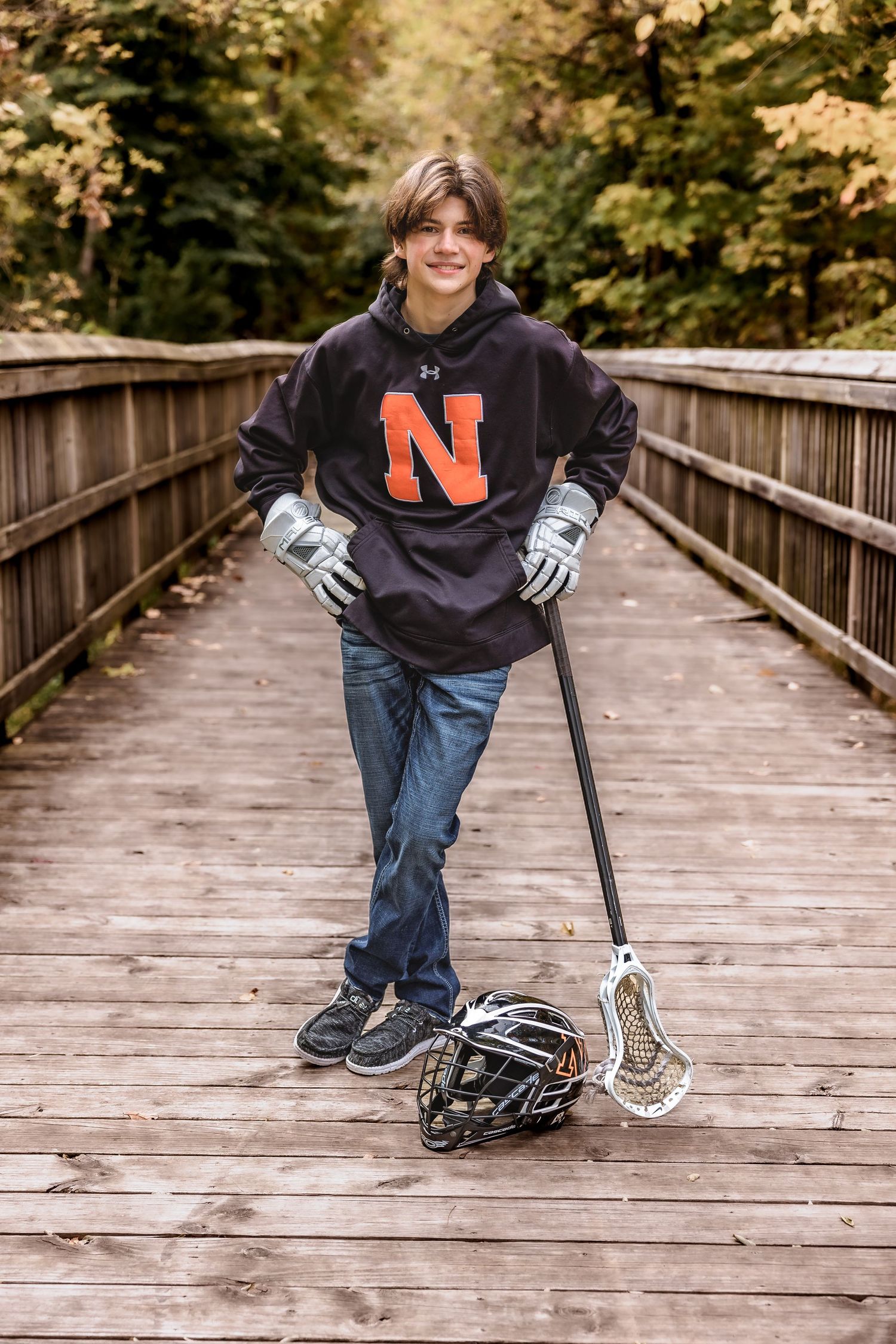 Student athlete in black 'N' sweatshirt holding lacrosse stick on wooden boardwalk.
