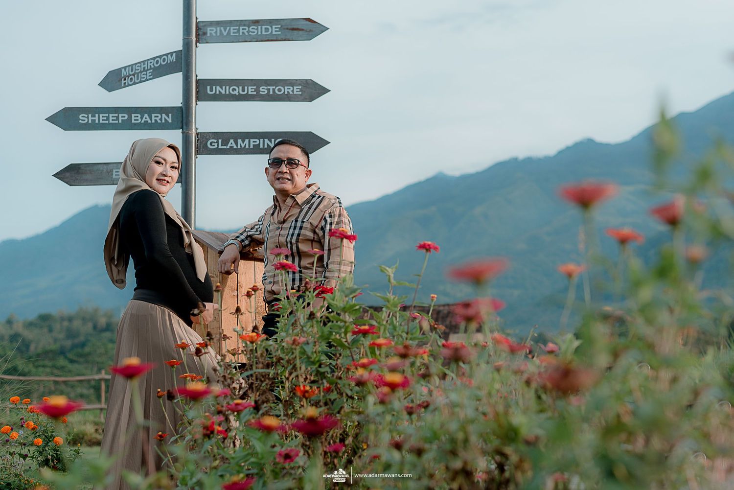 Couple near directional signs with red flowers and mountains in background.