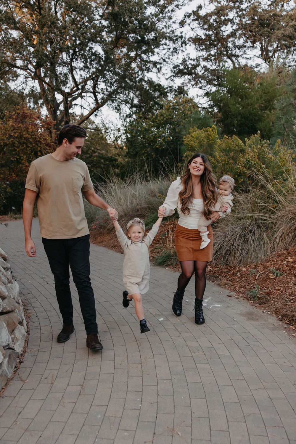 A playful family walks together on a brick path with children jumping and holding hands.