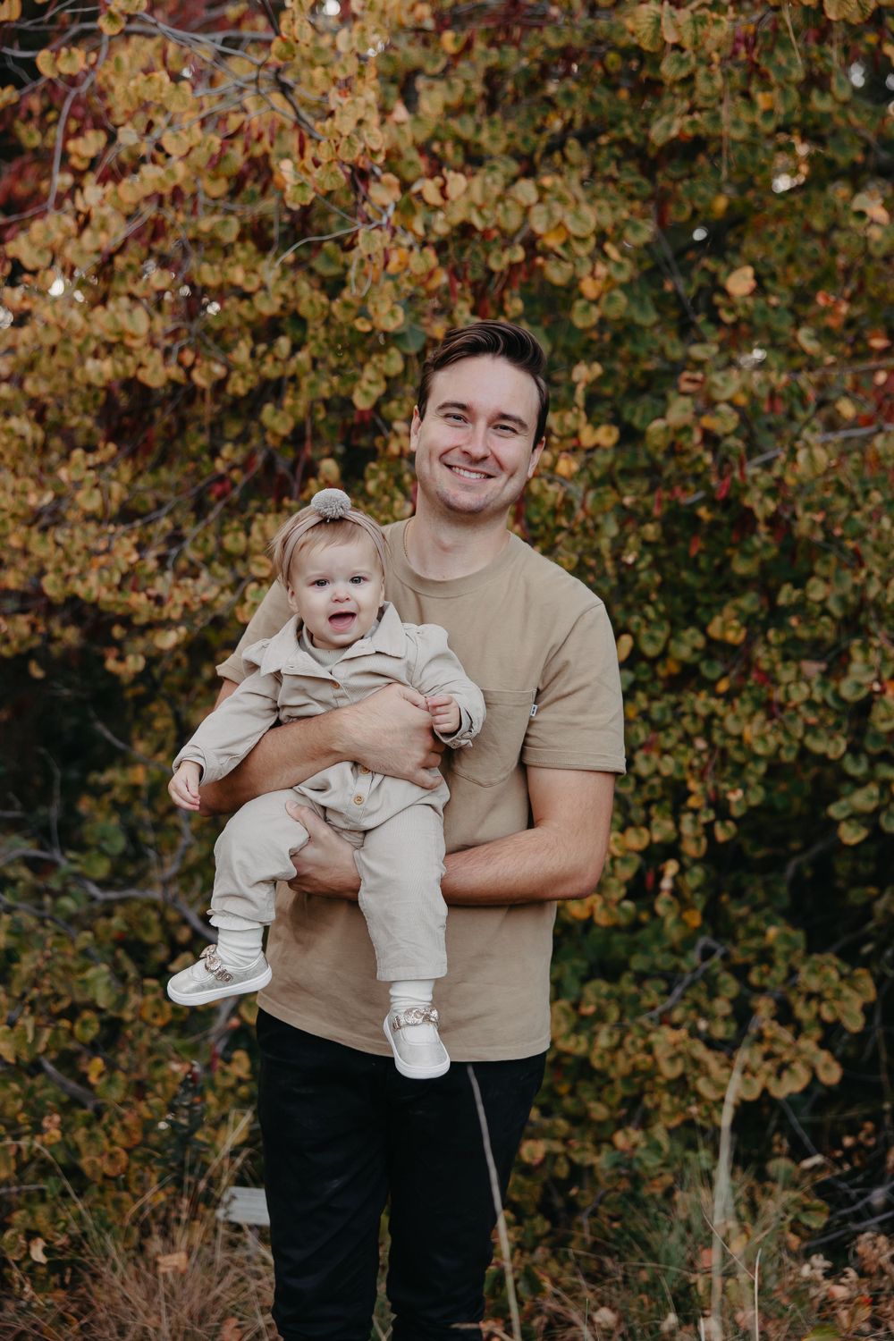 Parent holding an infant against autumn foliage backdrop in casual attire.