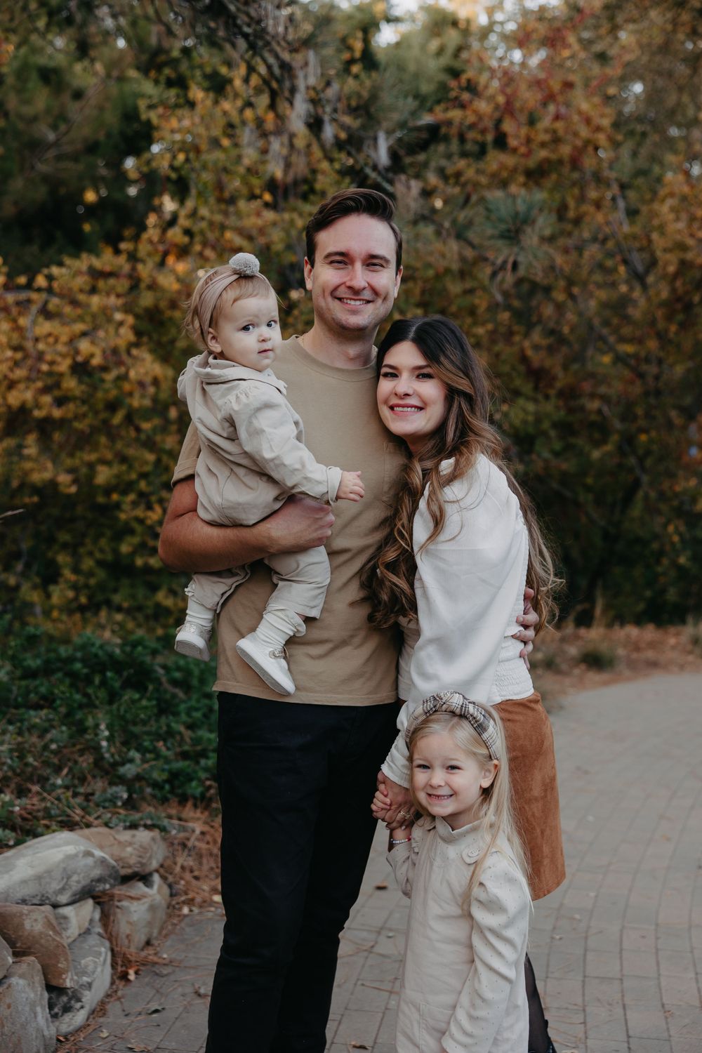 Family of four poses together on brick path surrounded by fall foliage in park setting.