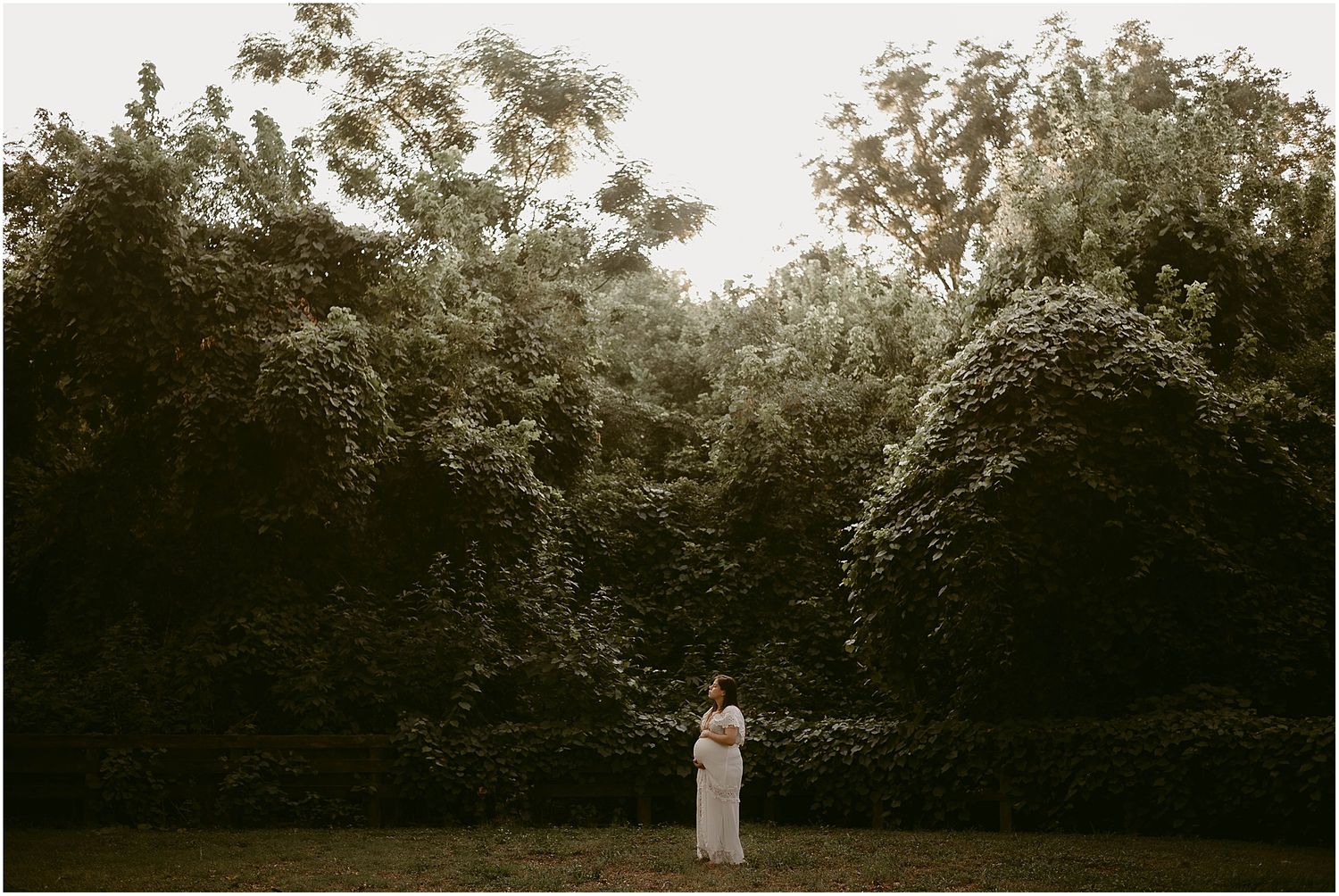 Wide shot of pregnant woman in white dress standing in garden surrounded by tall greenery.