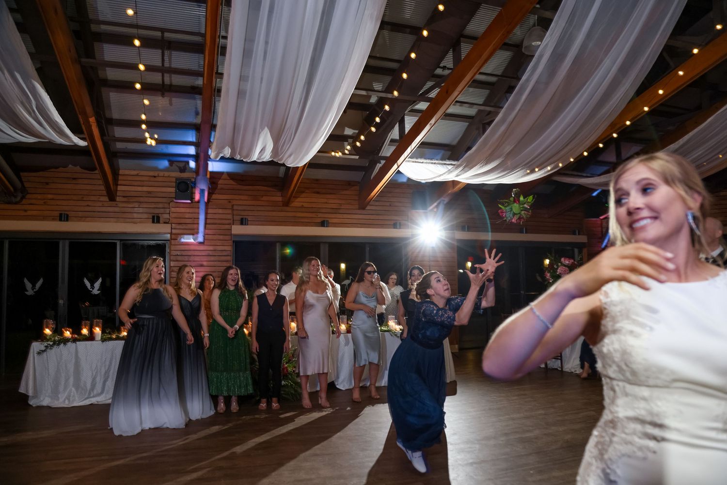 Wedding guests dancing and celebrating on a wooden dance floor under draped fabric and string lights at a reception venue.