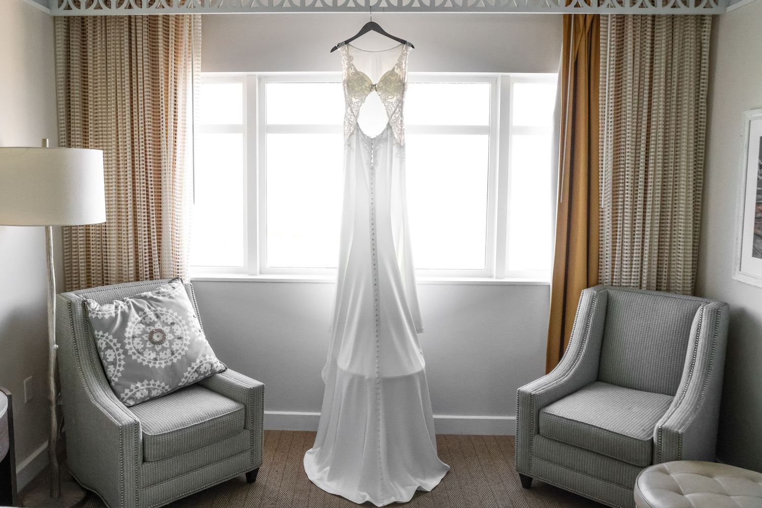 A white wedding gown hangs gracefully between two gray armchairs in front of a window with neutral curtains.