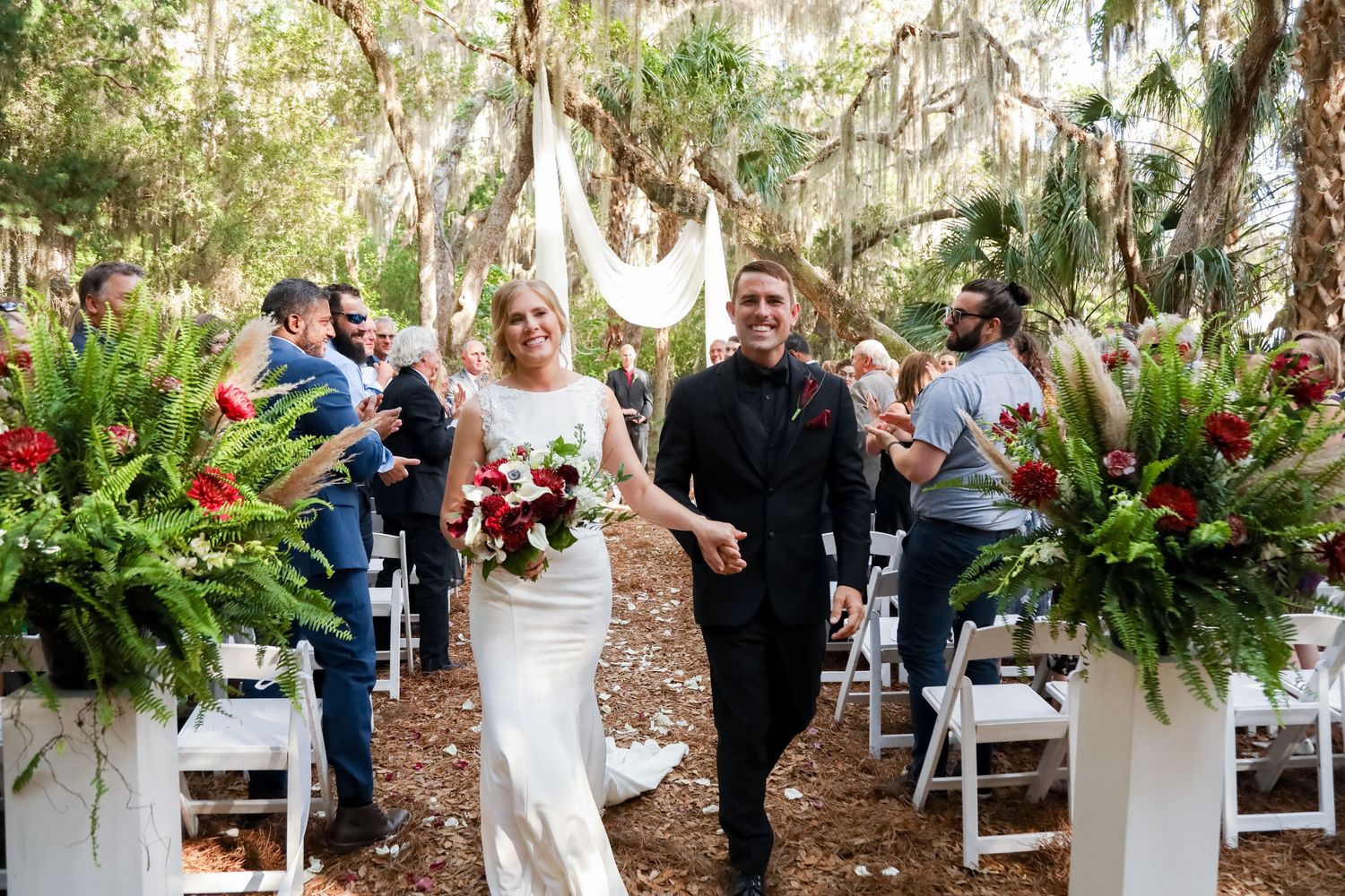 A joyful couple walks down an outdoor wedding aisle decorated with lush ferns under Spanish moss-draped trees.