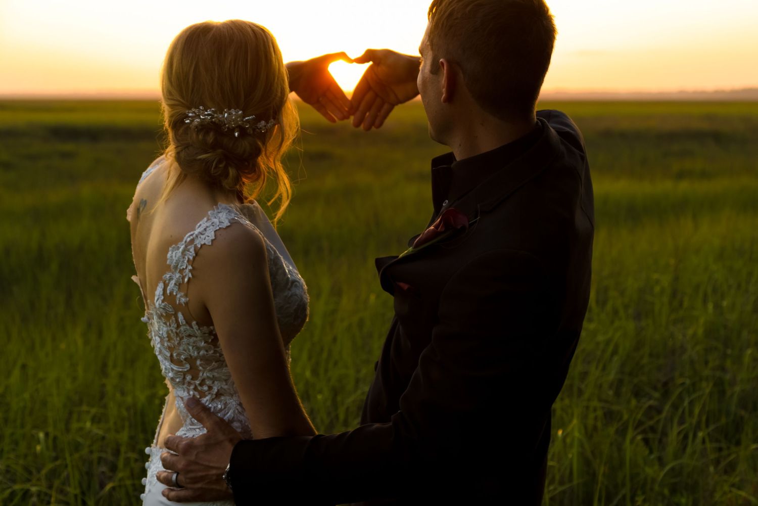 Two silhouetted figures make a heart shape with their hands against a golden sunset in a grassy field.