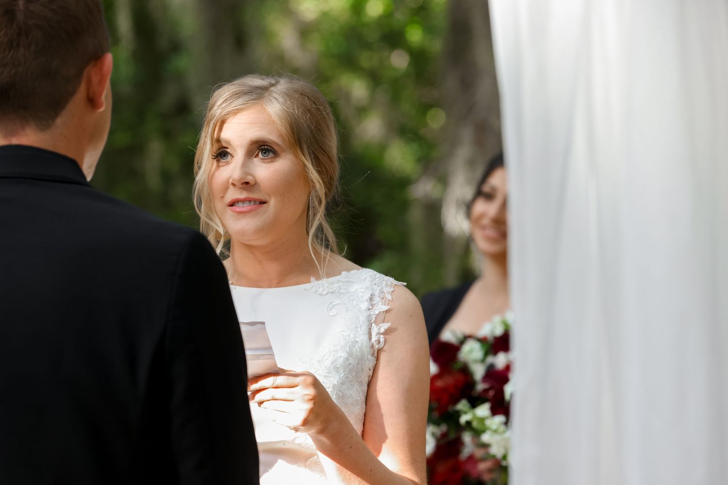 A couple exchanging vows during an outdoor wedding ceremony with floral decorations visible in the background.