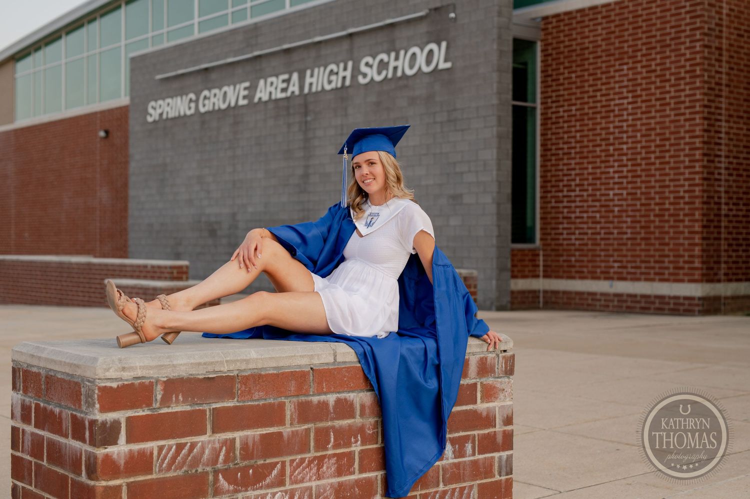 Graduate in blue cap and gown sits casually on brick ledge outside school building.