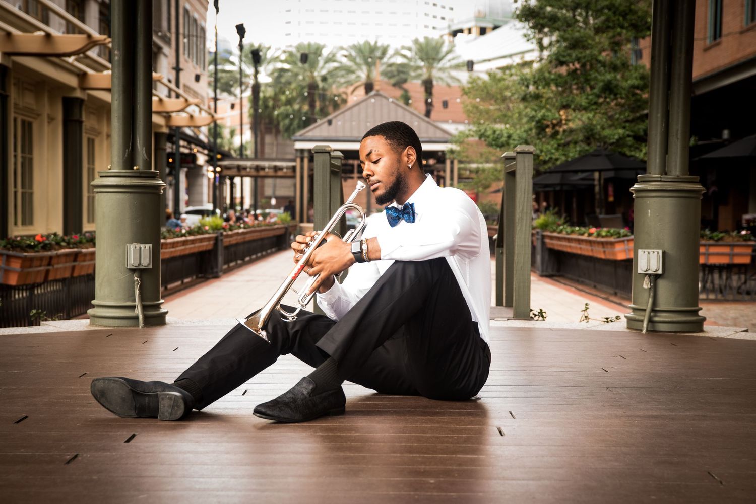 Graduate in formal attire sits on brick pavement in downtown setting with palm trees.