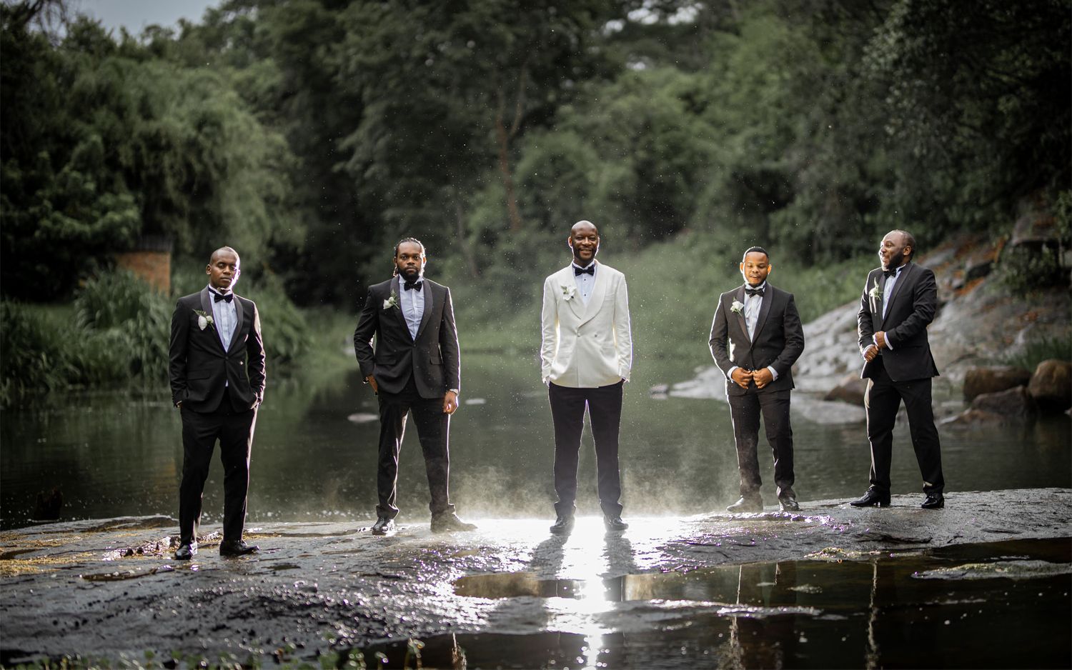 Groomsmen in formal attire standing on rocks by water creating a dramatic portrait.