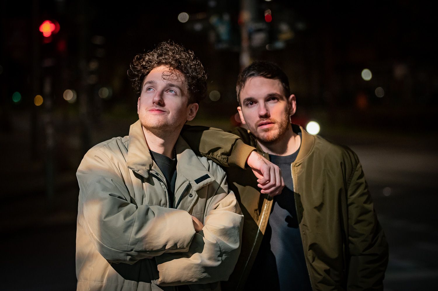 Two people posing on city street at night with urban lights in background.