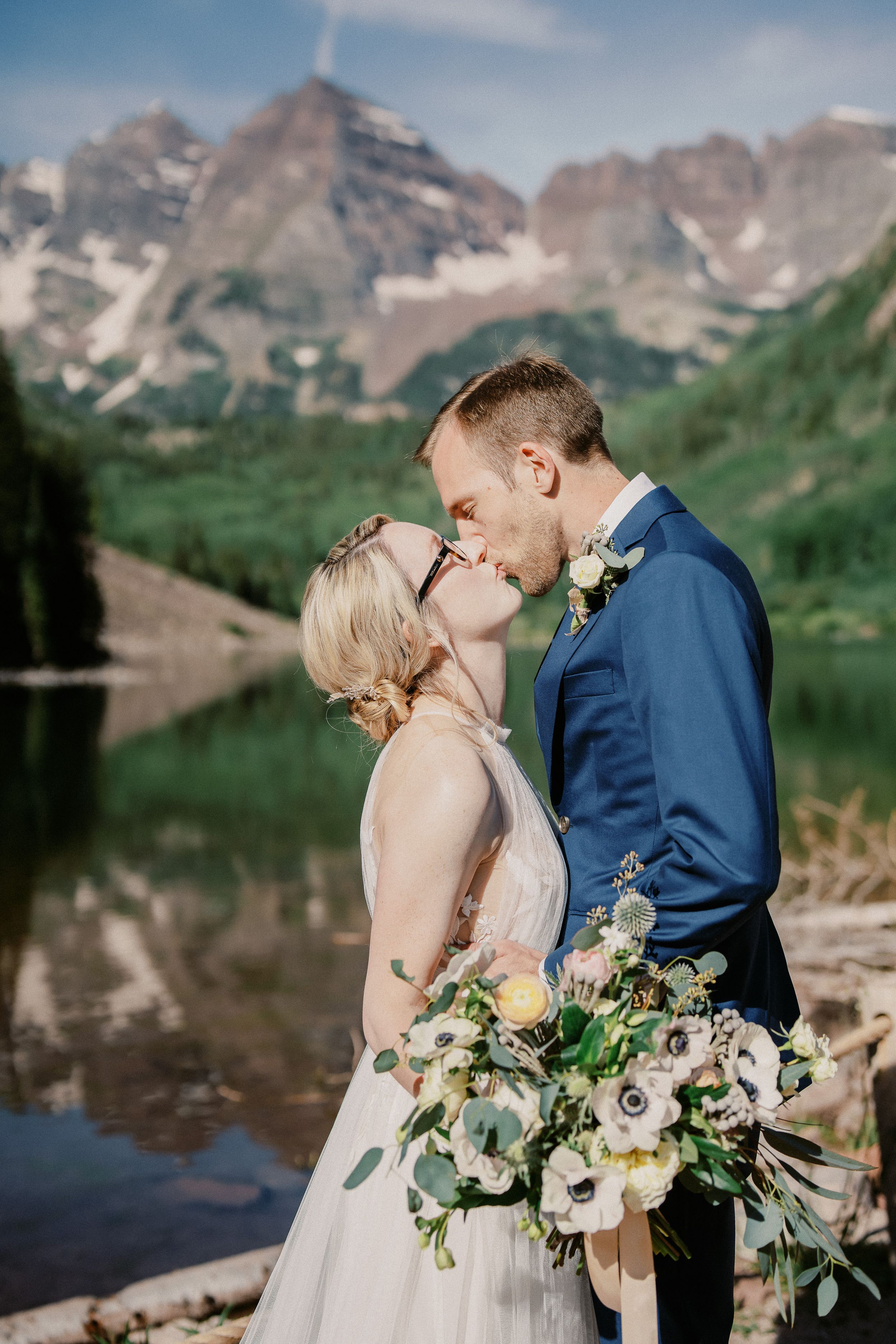 Bride and groom share intimate moment in front of majestic mountain lake landscape.
