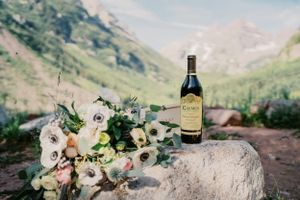Wedding bouquet with white anemone flowers and wine bottle against mountain backdrop.