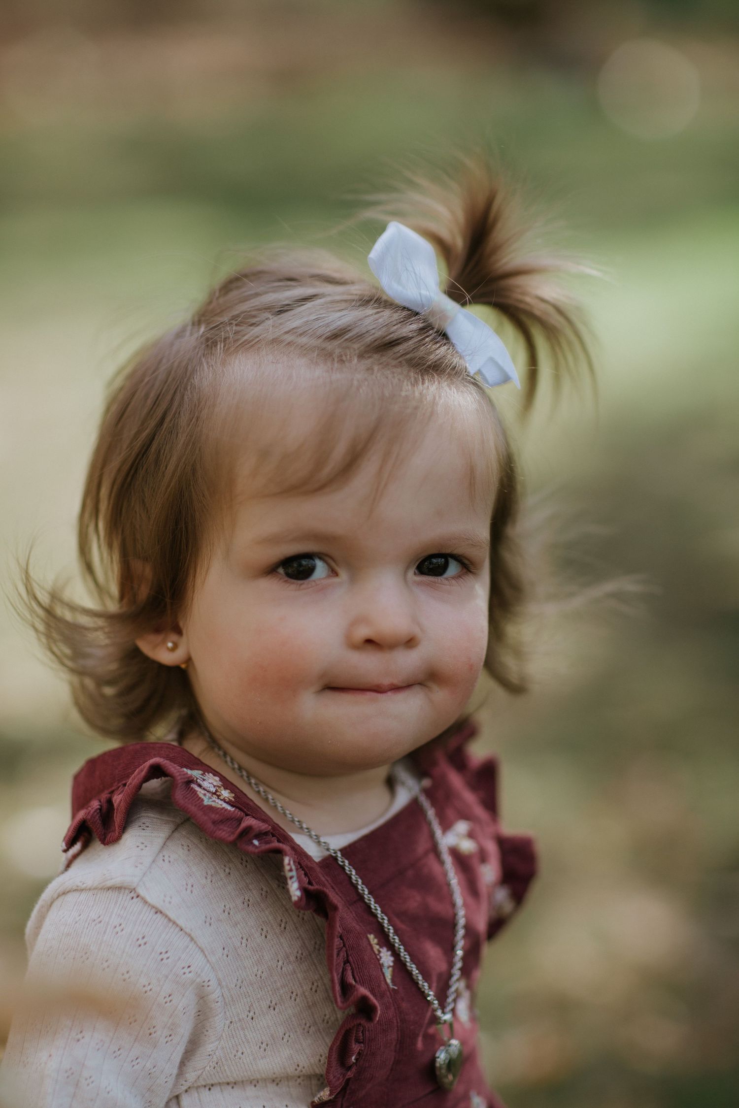 A sweet toddler with a blue bow wearing a cream sweater and burgundy vest smiles at the camera.
