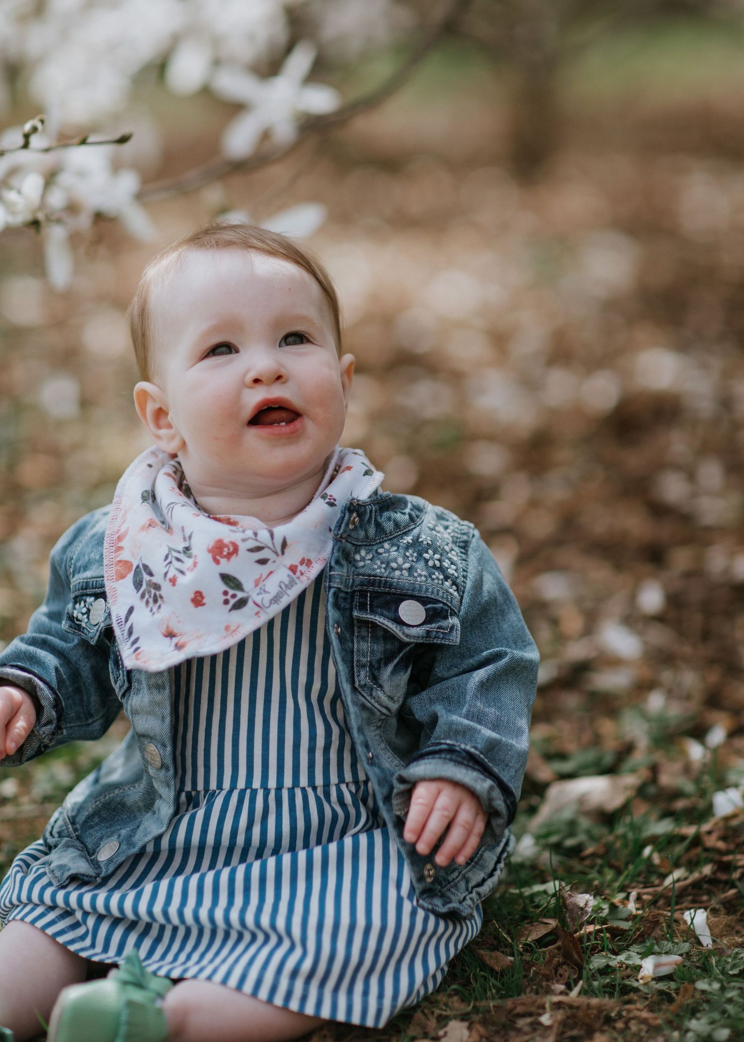 Baby wearing denim jacket and striped dress smiles while sitting under blooming tree.