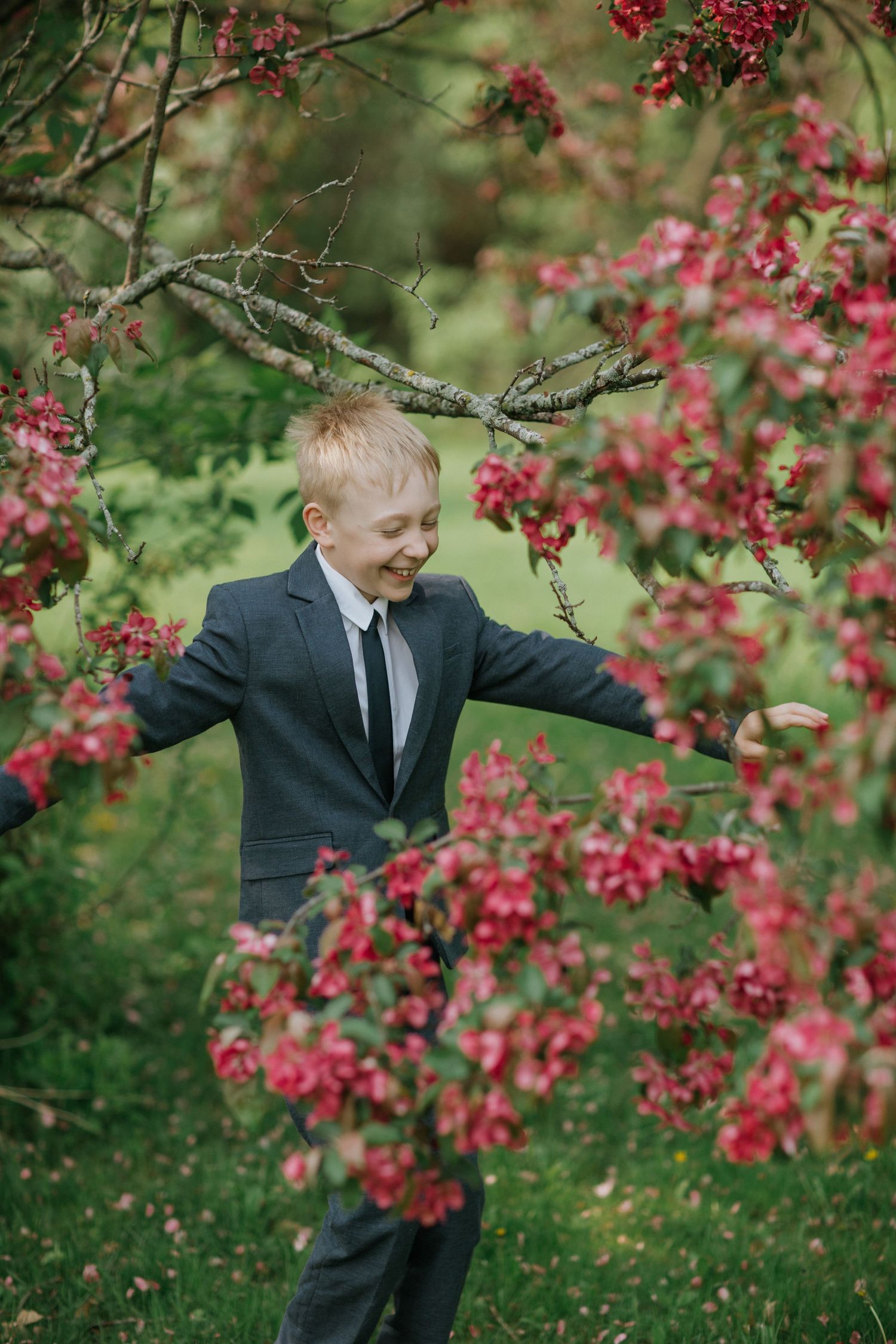 A young boy in a suit playfully poses among vibrant pink flowering branches.