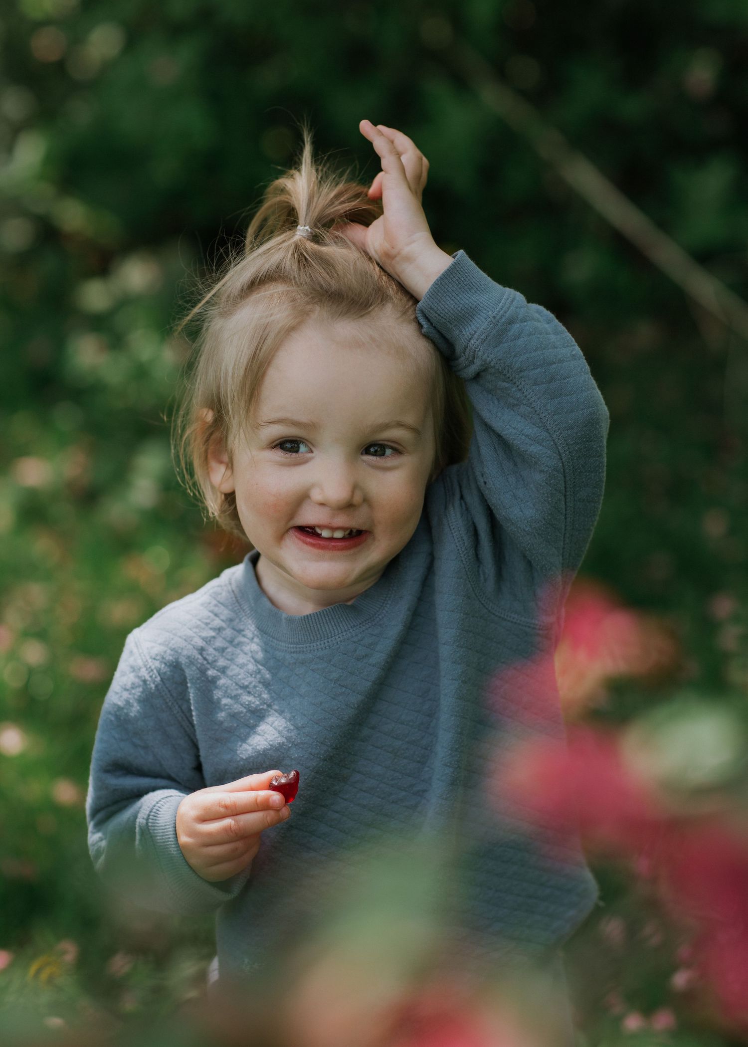 Toddler in grey sweater laughing and playing in garden with blurred flowers.