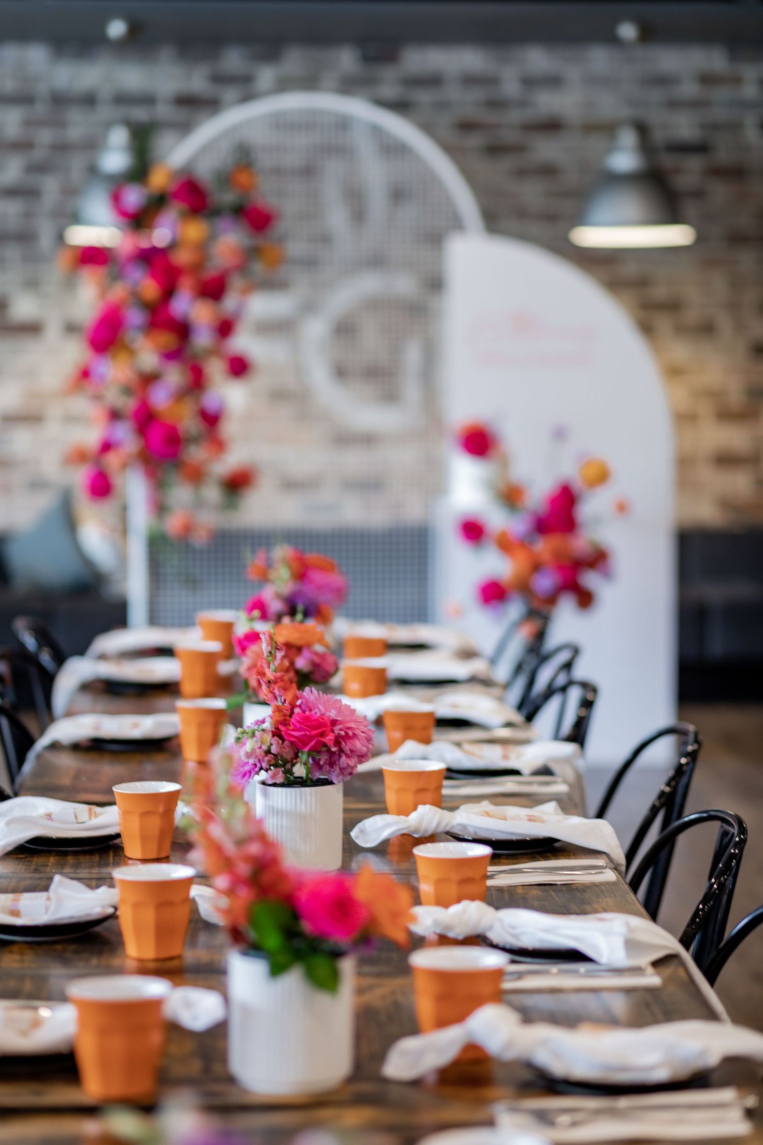 Elegantly set table with bright floral centerpieces, orange cups, and white mesh backdrop for special event.