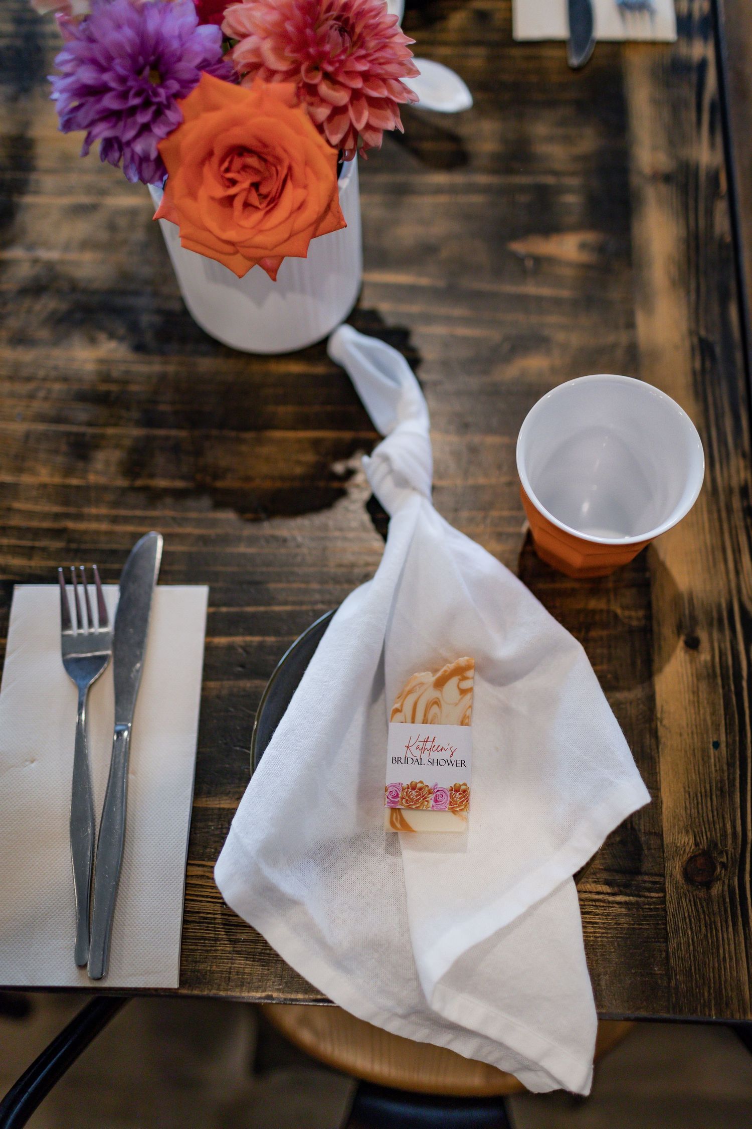 Table setting with orange and purple flowers in vase alongside white napkin on wooden surface.