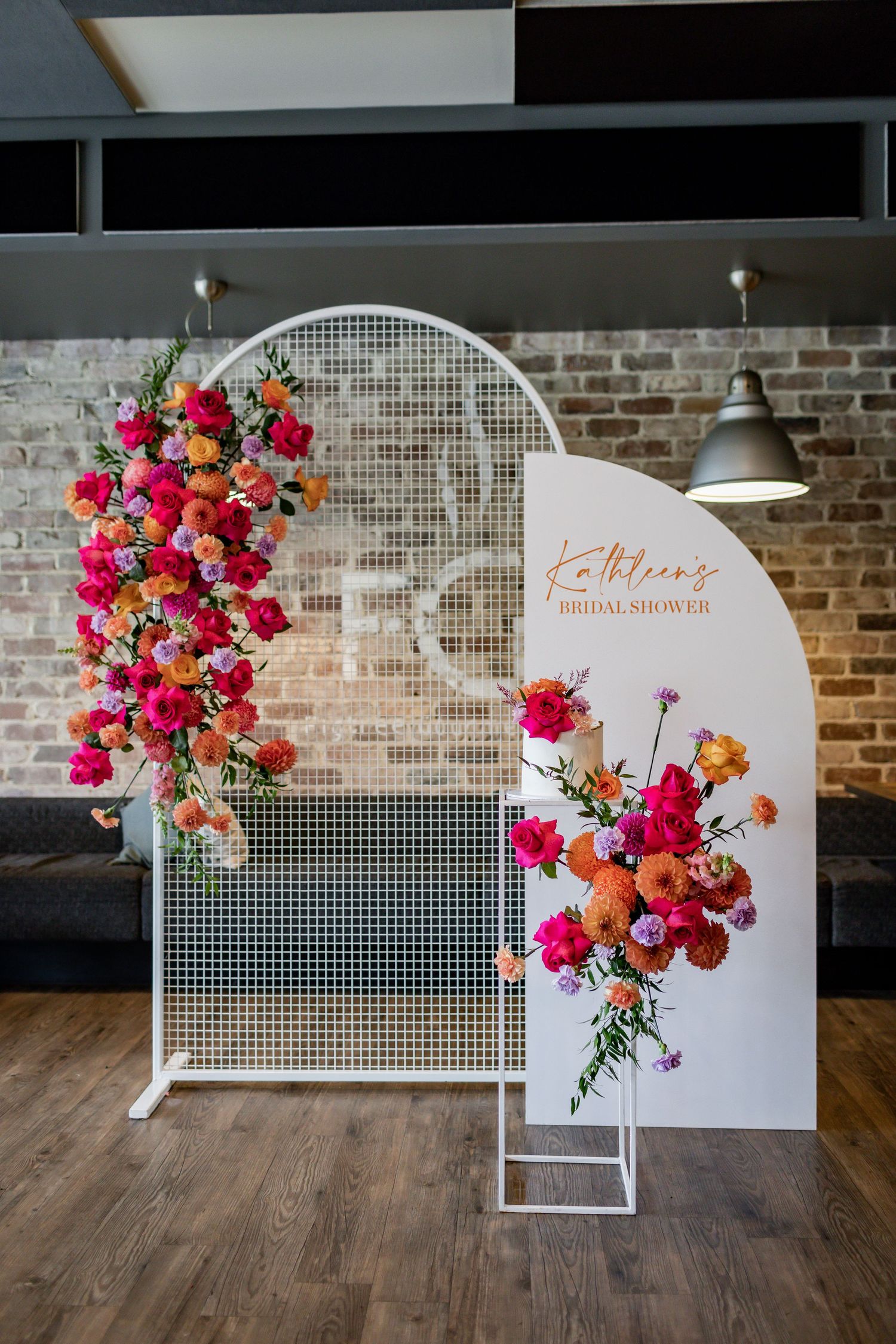 Modern bridal shower setup with white mesh backdrop and vibrant pink and orange floral arrangements against exposed brick wall.