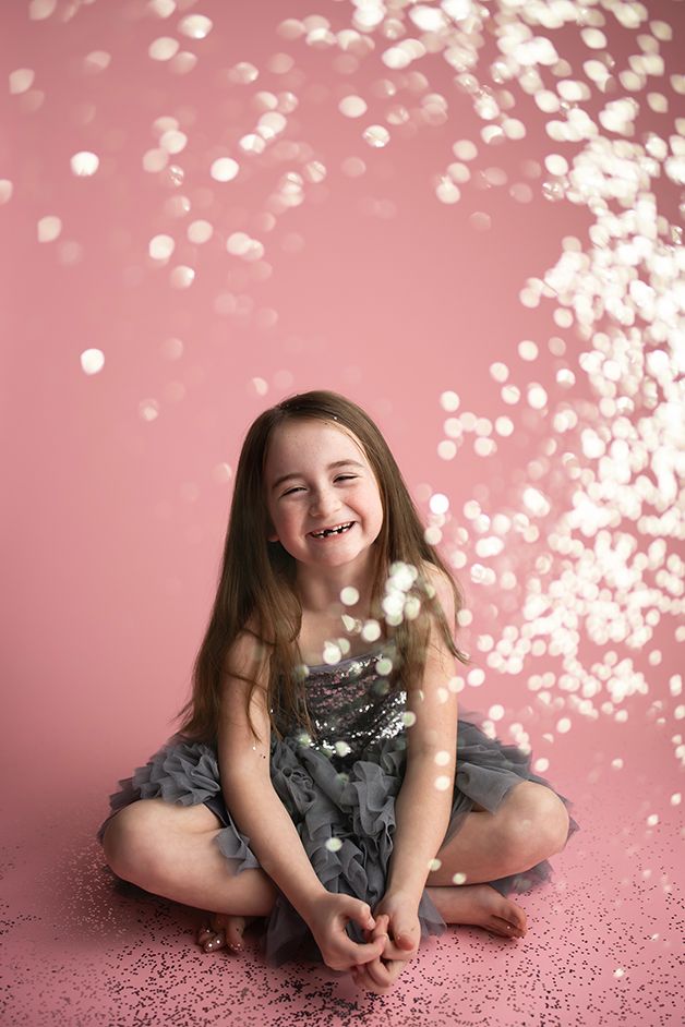 Little girl in grey dress sitting and laughing with silver glitter falling around her.