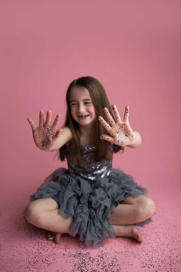 Little girl sitting on pink backdrop laughing and showing the glitter that is stuck to her hands.