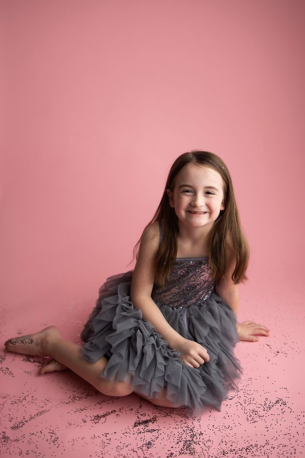Little girl in grey dress sitting on pink backdrop with silver glitter scattered on the ground.