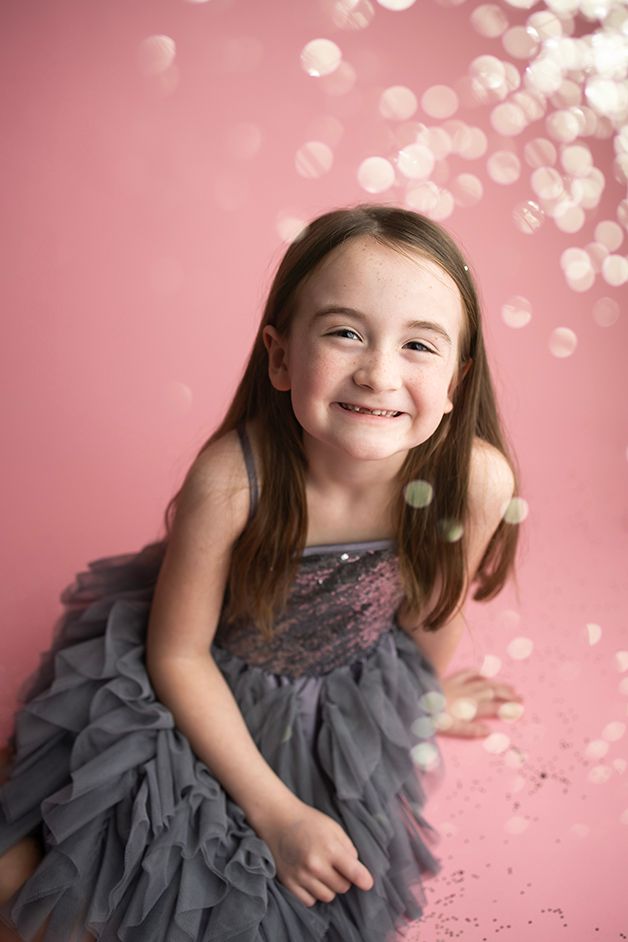 Little girl sitting on pink backdrop smiling at the camera with silver glitter falling.