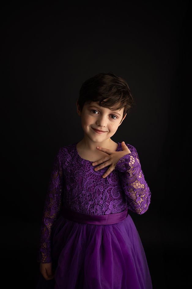 Little boy in purple dress standing on black backdrop smiling towards the camera.