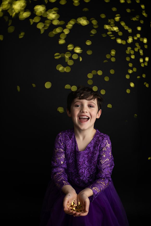 Little boy in a purple dress on a black backdrop throwing gold glitter.