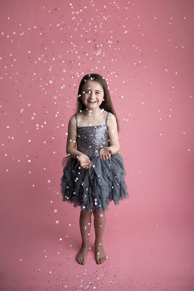 Child standing in a grey dress on a pink backdrop with silver glitter falling.