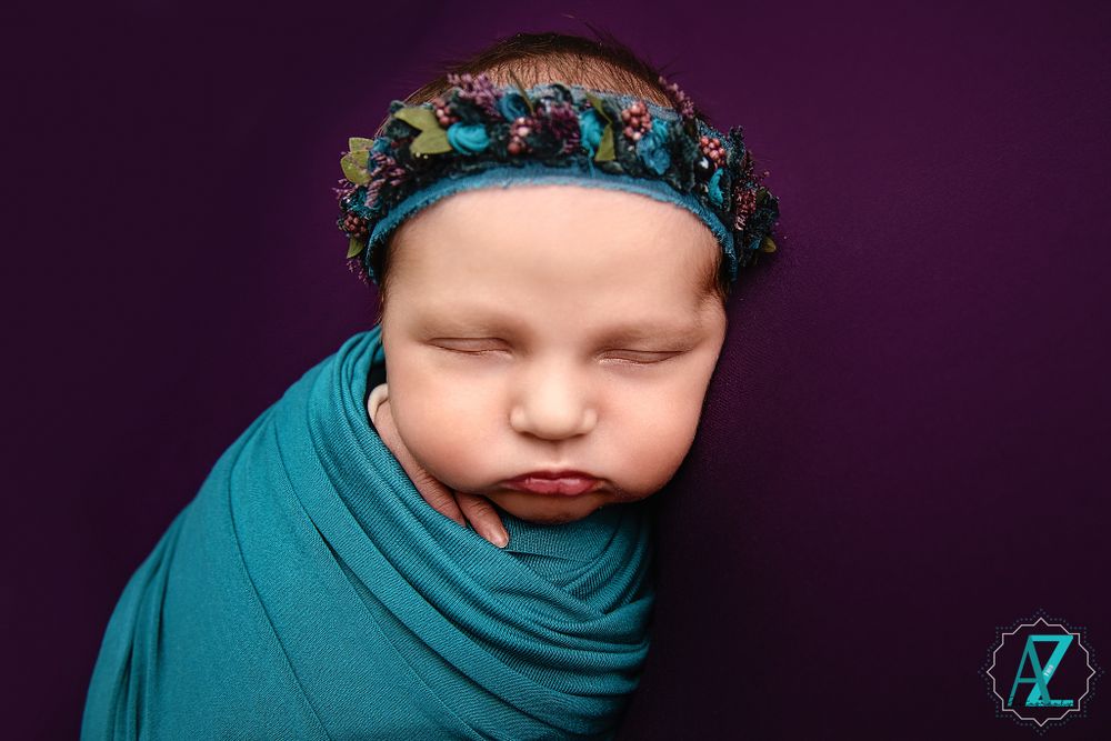 Sleeping baby wrapped in teal fabric wearing turquoise floral crown against purple background.