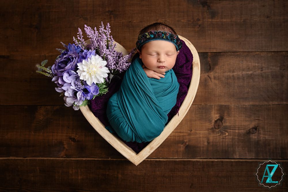 Heart-shaped basket holds newborn with teal wrap beside purple floral arrangement.