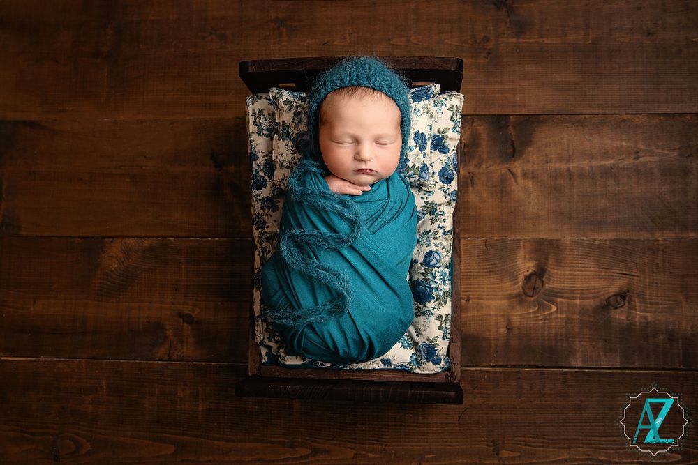 Swaddled newborn rests in floral patterned bed on wooden background.
