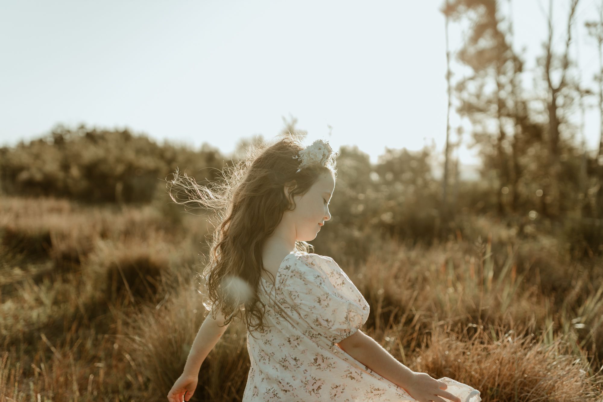 A figure in a white dress spins joyfully in a sunlit field with grass swaying in the breeze.