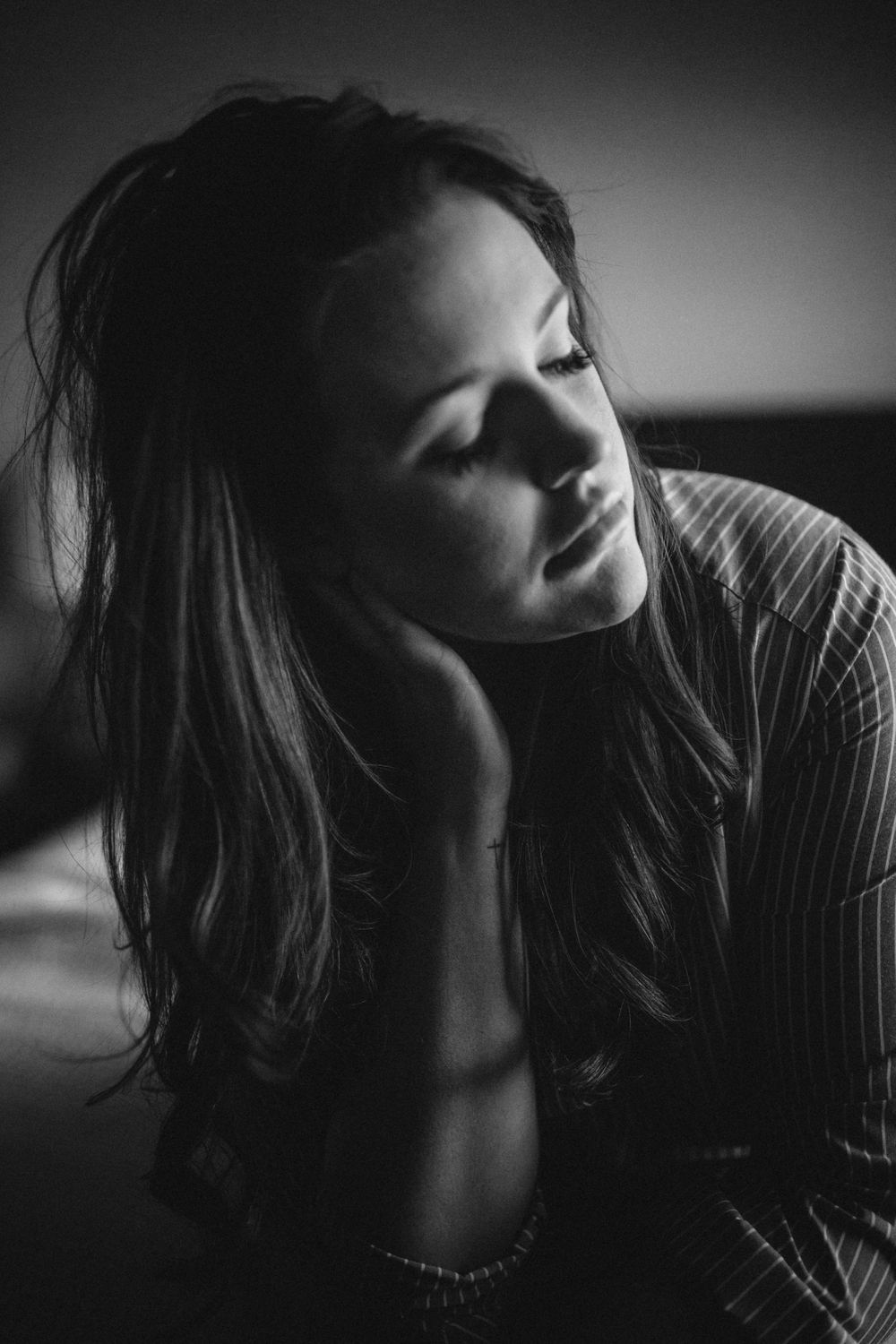 black and white image of woman sitting on bed, in dark natural window light, eyes closed, hand on neck
