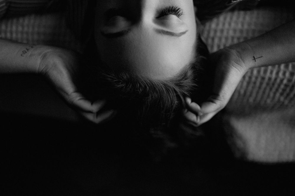 black and white image of woman laying back on bed, hands in hair, from the nose up
